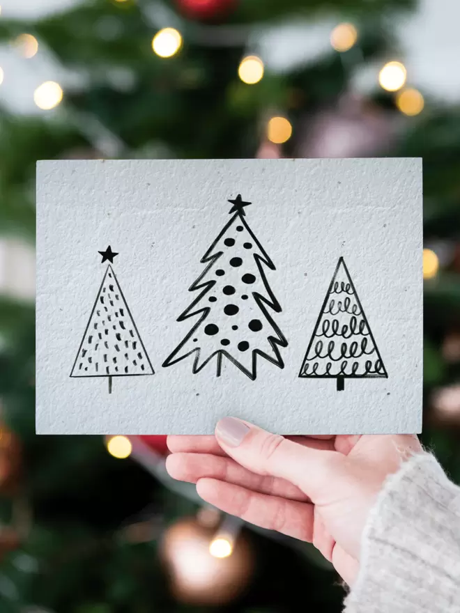 A hand holding a card with three black and white illustrated Christmas trees with a blurred tree and lights in the background.