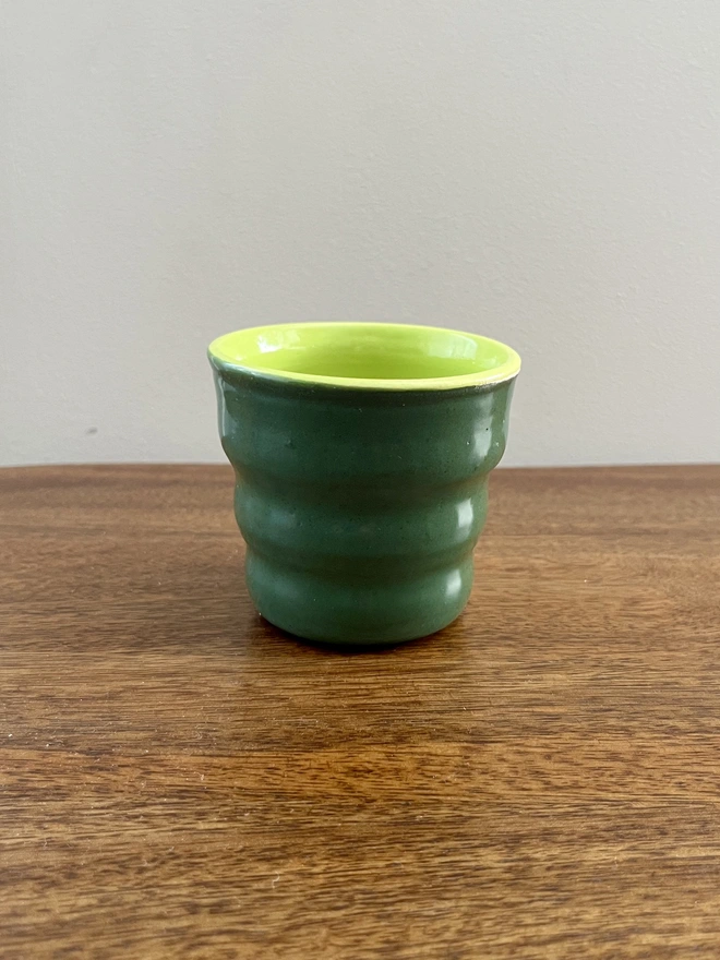mini ceramic tumbler with wavy sides on a wooden table. the tumbler has a forest green glaze on the outside and lime green on the inside and rim. 
