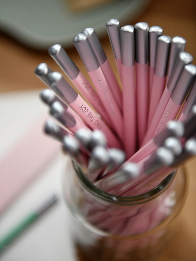 A jar of pink pencils stands on a wooden desk surrounded by stationery items. Each pencil has the words Find The Joy on the side.