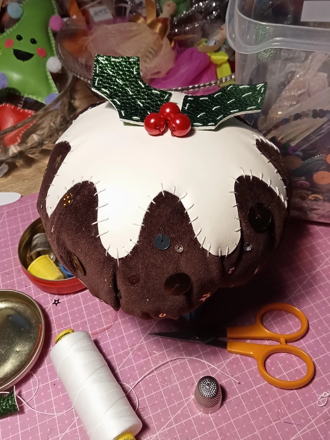 A plump velvet Christmas pudding decoration sits on a pink cutting mat surrounded by thimble, thread, scissors and other sewing paraphernalia