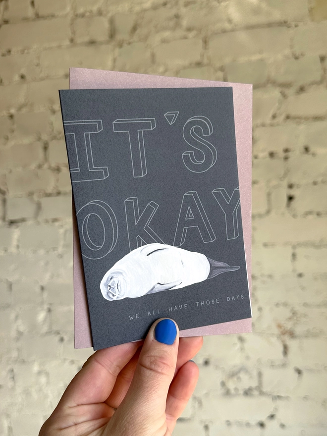 a dark grey greetings card featuring an illustration of a seal with the text “It’s Okay - we all have those days” with a muted purple envelope