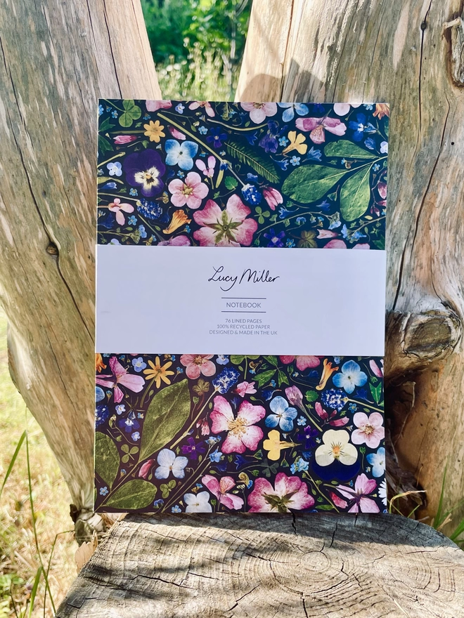 Eco-friendly Notebook with Pressed Flower Cover, Nature-Inspired, 'Lucy Miller' Branded Belly Band, Leaning Against Tree Trunk.