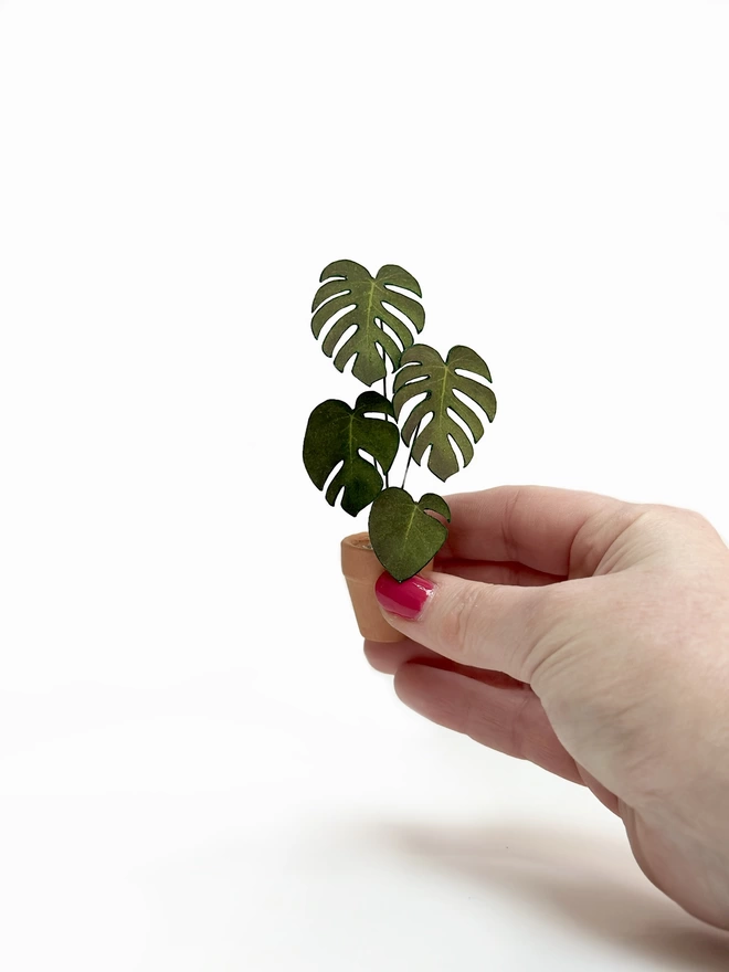 A miniature replica Monstera Deliciosa paper plant ornament in a terracotta pot held between 3 fingers against a white background