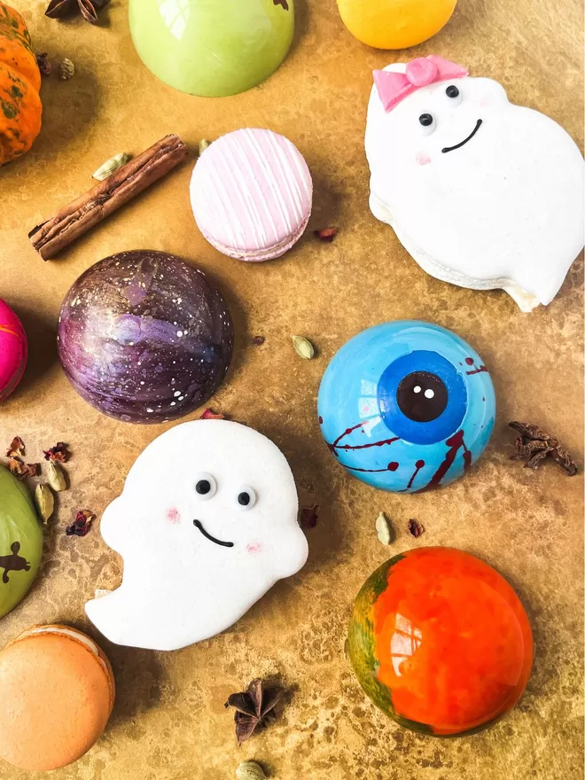 colourful halloween macarons, ghost macarons and halloween hot chocolate bombes featuring a bloodshot eye and a frankenstiens monster design on a gold background with little pumpkins