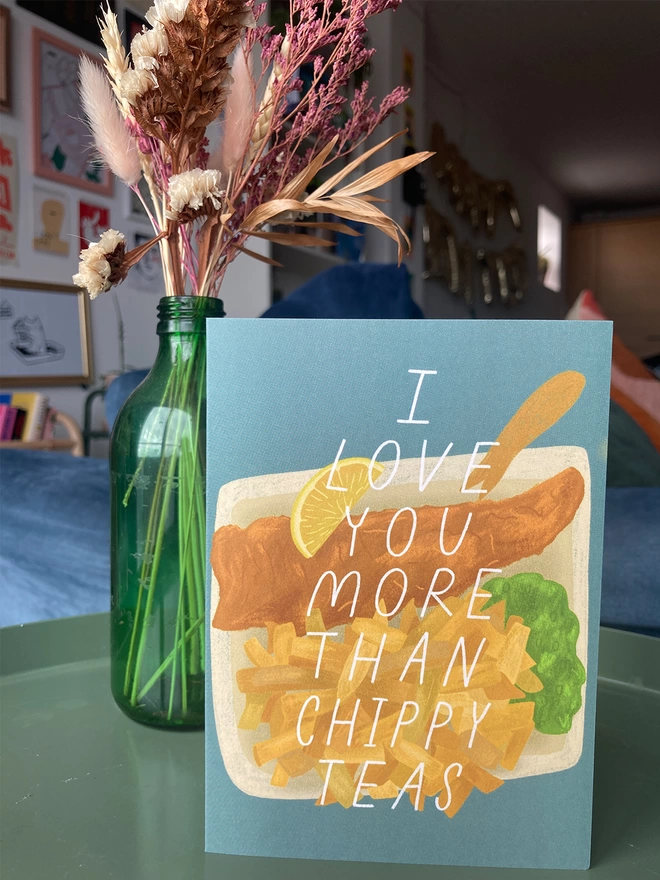 Card saying 'I love you more than chippy teas', placed on table with flowers in the background.