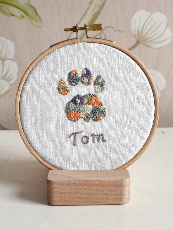Sunshine Garden Cat Paw of Golden Yellows and Bright Orange Blossoms with Green French Knot grass background.  Displayed in an embroidery hoop on a wooden stand. 