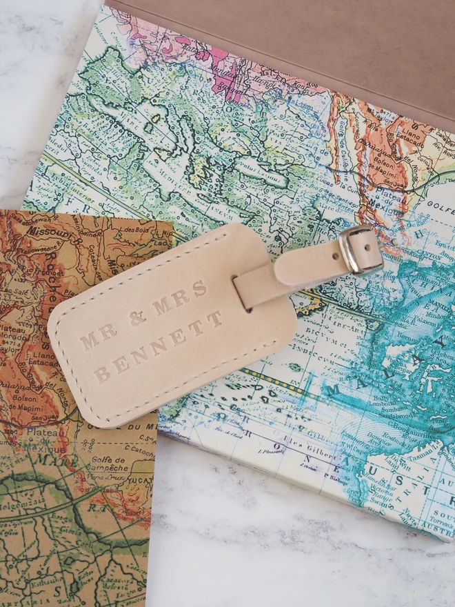 Personalised Leather Luggage Tag. A natural cream leather luggage tag, personalised with a newly married couples names. Sitting on a collection of colourful maps.