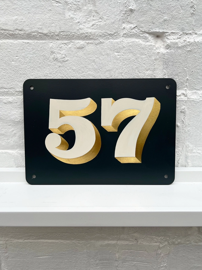 Hand painted house number against a white brick wall, number 57 in off white and directionally burnished 23 carat gold leaf. 