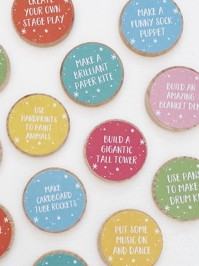 Several wooden tokens with brightly coloured labels, each with a fun activity idea printed on, lay on a white surface.