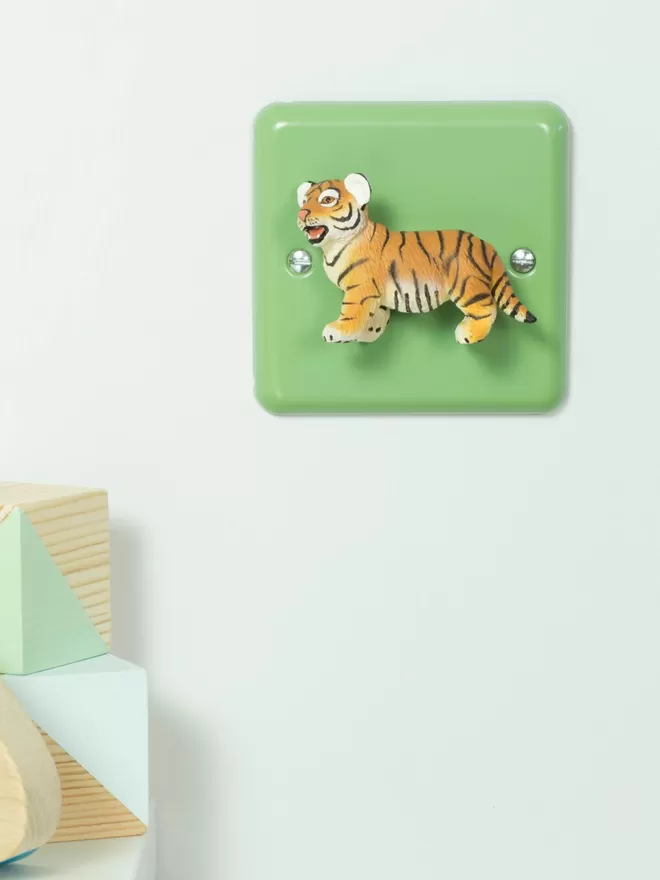 A tiger light switch on a white wall with wooden nursery building blocks sitting on white wooden shelf to the left. The light switch is a pastel green nursery dimmer light switch with a tiger as the rotary knob to turn the lights on and off. The light switch plate is beryl green and made of metal, epoxy coated steel by Varilight. The tiger is made of plastic. The animal light switch brand is Candy Queen Designs.