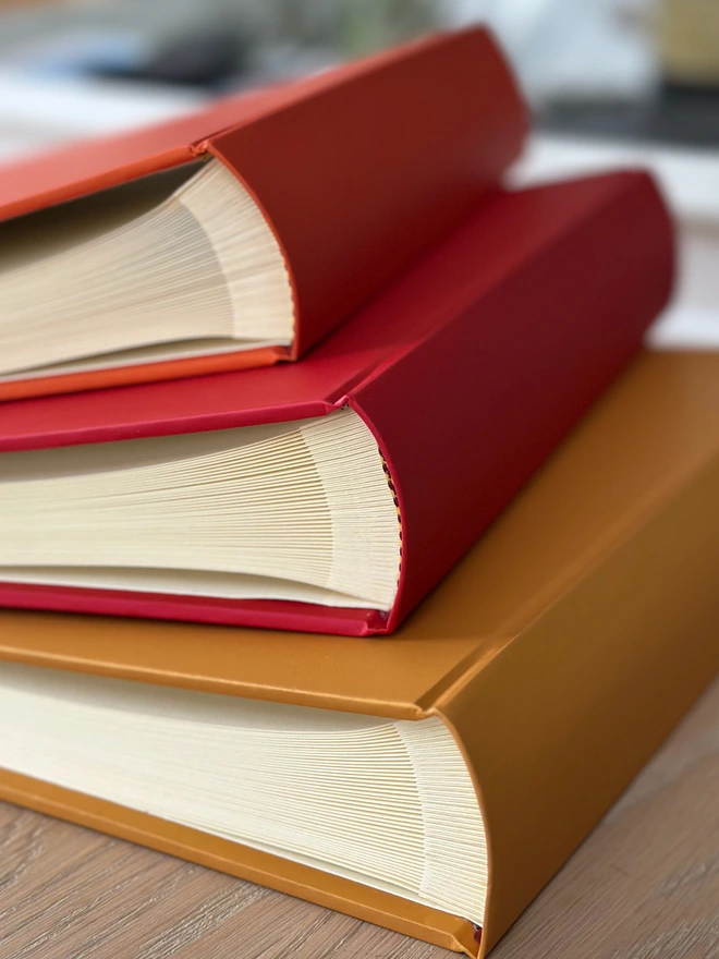 A pile of hardback photo albums rests on a wooden surface. The colours are orange, red and yellow.