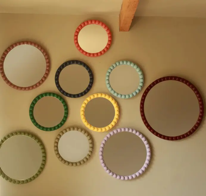 Many round oversized bobbin mirrors, in different colours and sizes with a decorative bobbin-style wooden frame, hanging on a white wall.