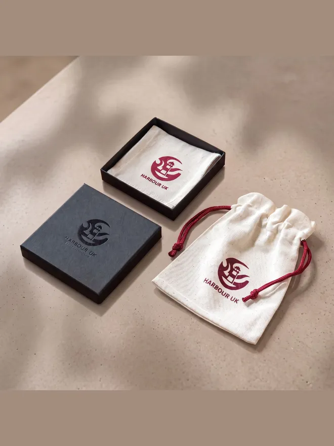 Harbour UK packaging: an open gift box with a cream logo pouch inside, a closed charcoal gift box, and a cream drawstring pouch with a maroon cord, all printed with the Harbour UK lighthouse logo.