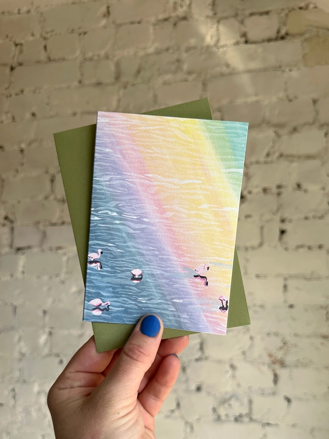 a greetings card featuring an illustration of flamingos in water with a rainbow reflection with a green envelope 