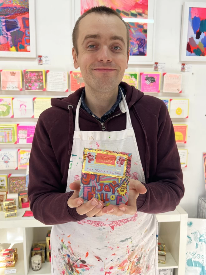 happy artist holding a bright coloured packaging of charity full of joy ruby chocolate buttons
