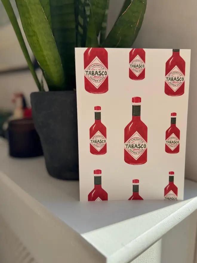 tabasco bottles illustrated card. card photographed on mantelpiece with houseplant in the background.