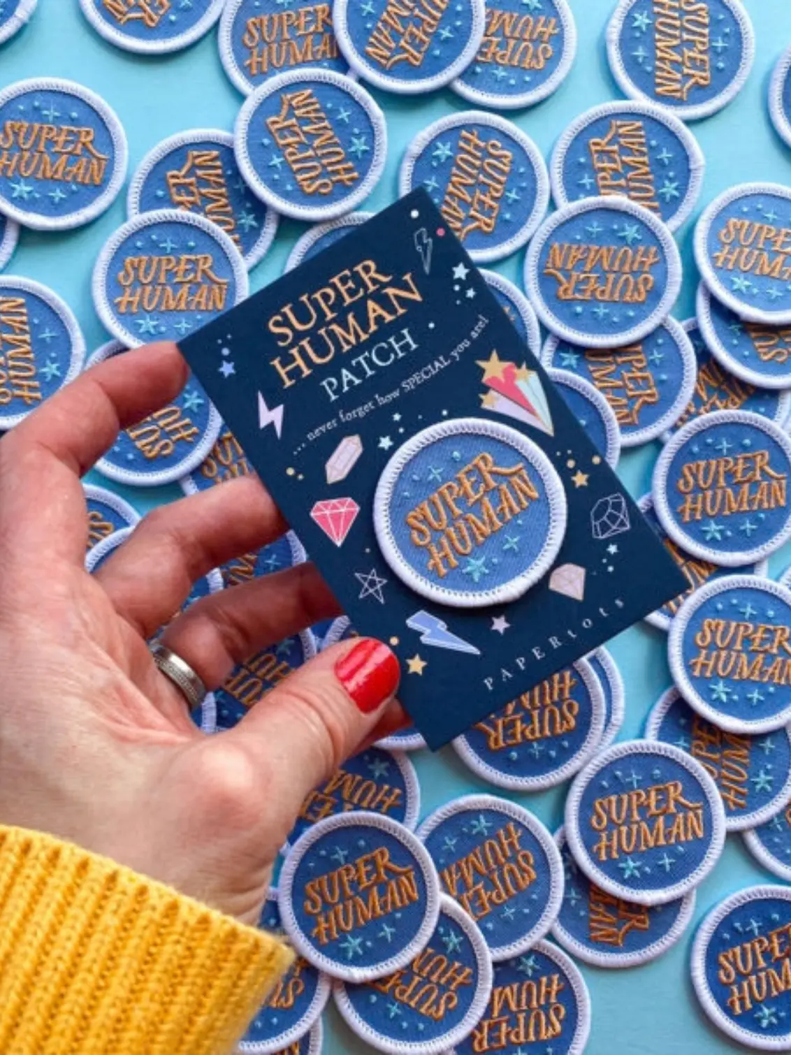 A hand holds a navy "Super Human Patch" card with a round embroidered patch in blue and peach. Dozens of matching patches are scattered across a pale blue surface below.