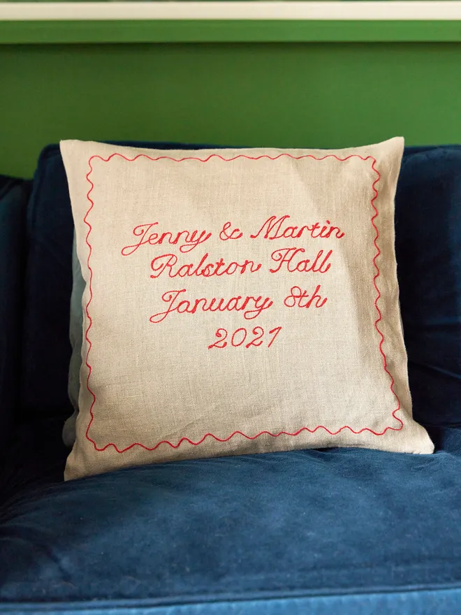 Embroidered linen cushion with red stitching for a wedding anniversary and date, on a blue sofa with a light blue cushion behind, against a bright green wall