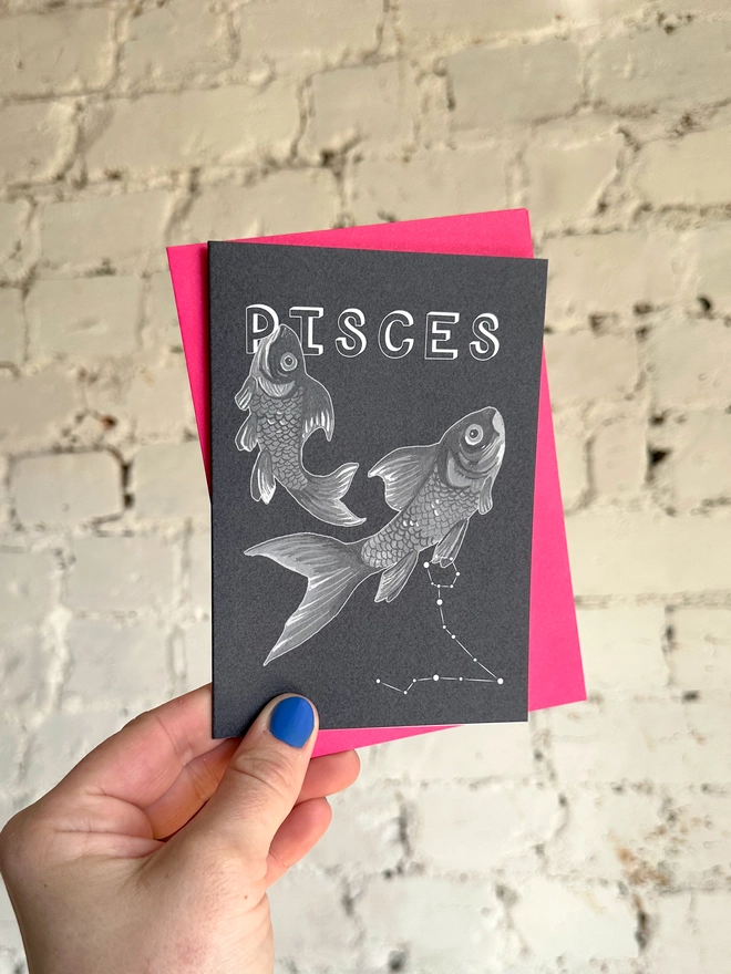 a greetings card featuring a greyscale illustration of a zodiac style couple of fish with the text “PISCES” with a neon pink envelope