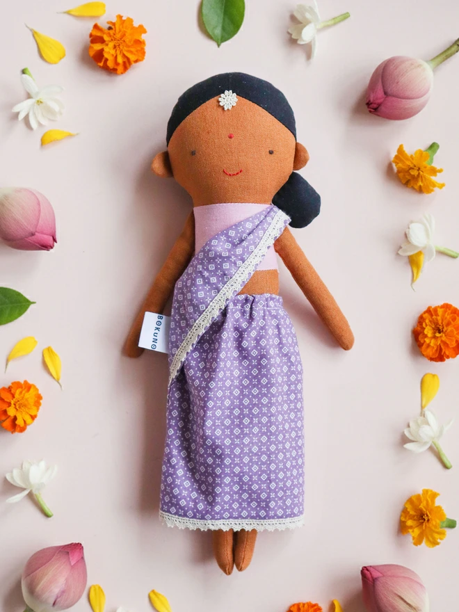 skin tone, wearing a lilac patterned sari with lace trim and a pink top. The doll has black hair styled in a low side bun with a small white flower accessory on her forehead, simple stitched facial features. 