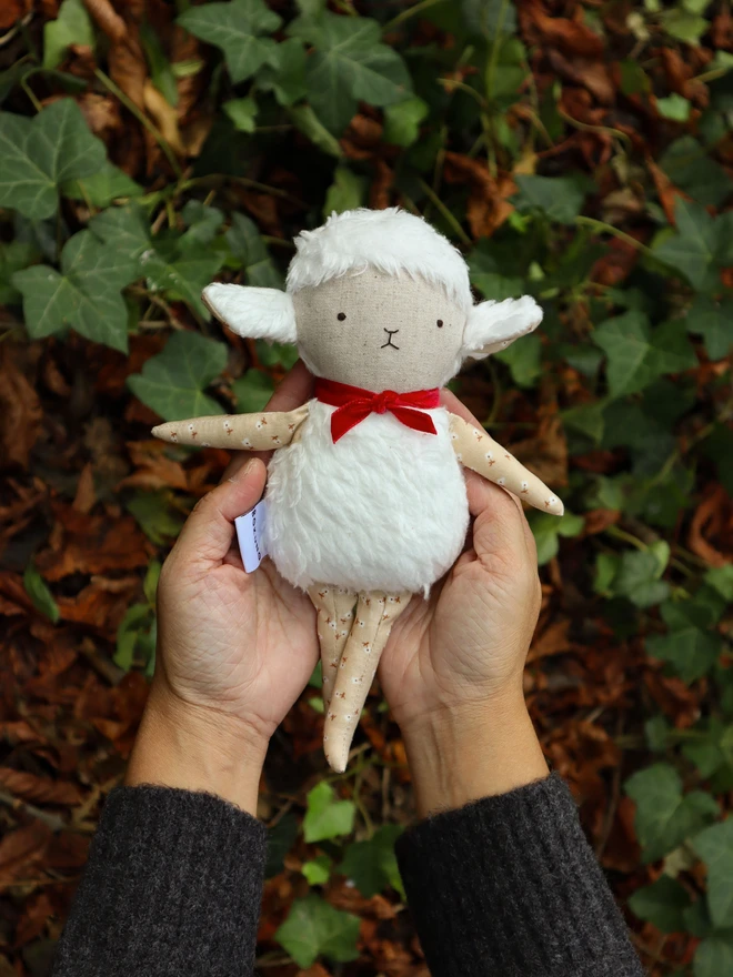 Handmade lamb doll with a fluffy white body, stitched face, beige floral fabric arms and legs, and a small red velvet bow tied around its neck.