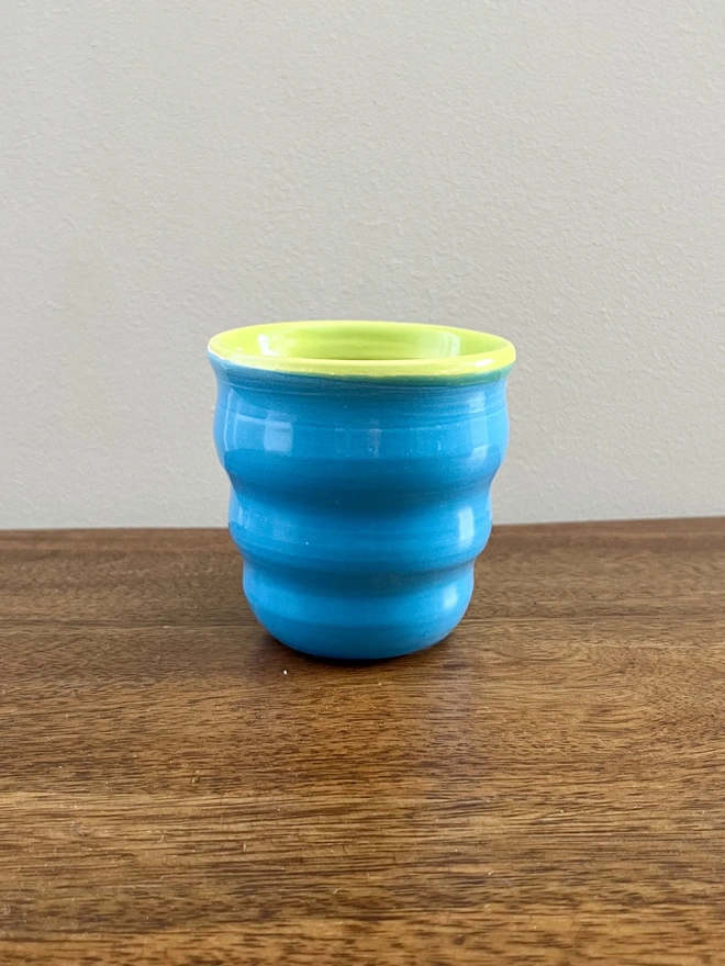 mini ceramic tumbler with wavy sides on a wooden table. the tumbler has a azure blue glaze on the outside and lime green on the inside and rim. 