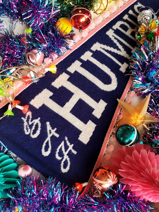 An image showing a table full of brightly coloured retro Christmas decorations. Amongst the tinsel, coloured lights, glass baubles and paper christmas trees is a navy blue knitted pennant flag that reads Bah Humbug in white sparkly letters.