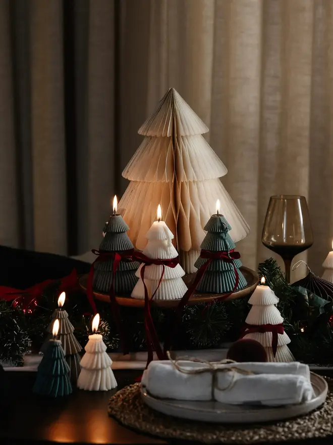 Tiered Christmas Tree Candle. Lit Christmas tree candles in cream, deep green and white, some tied with burgundy velvet bows, grouped around a paper tree on a festive table. The mix of colourways creates a striking, modern holiday display.