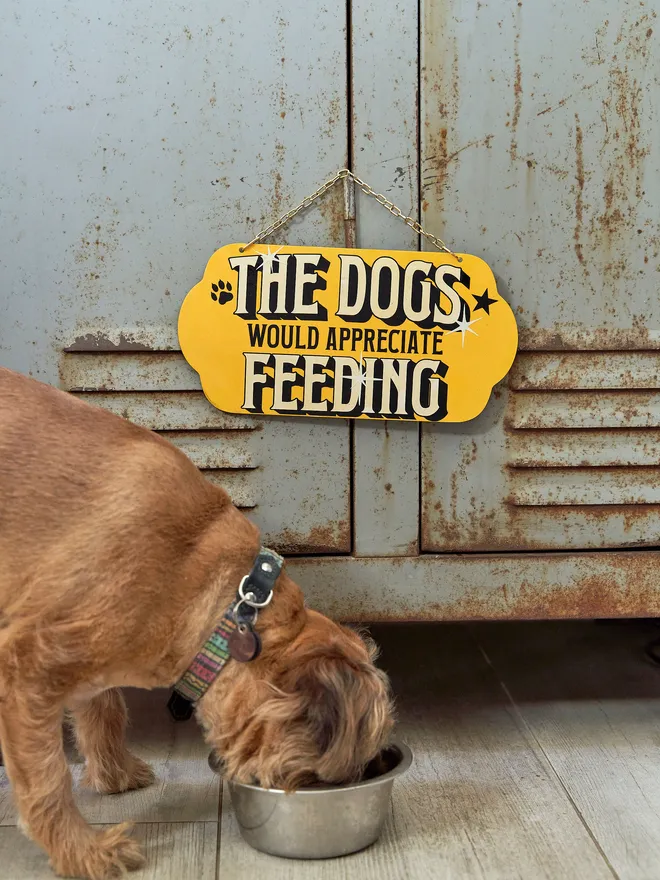 'The Dogs Would Appreciate Feeding' hanging sign with a gold chain, yellow background with white writing and paw prints on 