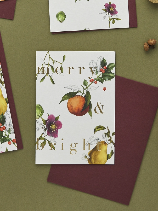 ivory based christmas card with festive hand drawn botanicals with gold foil wording and a burgundy envelope