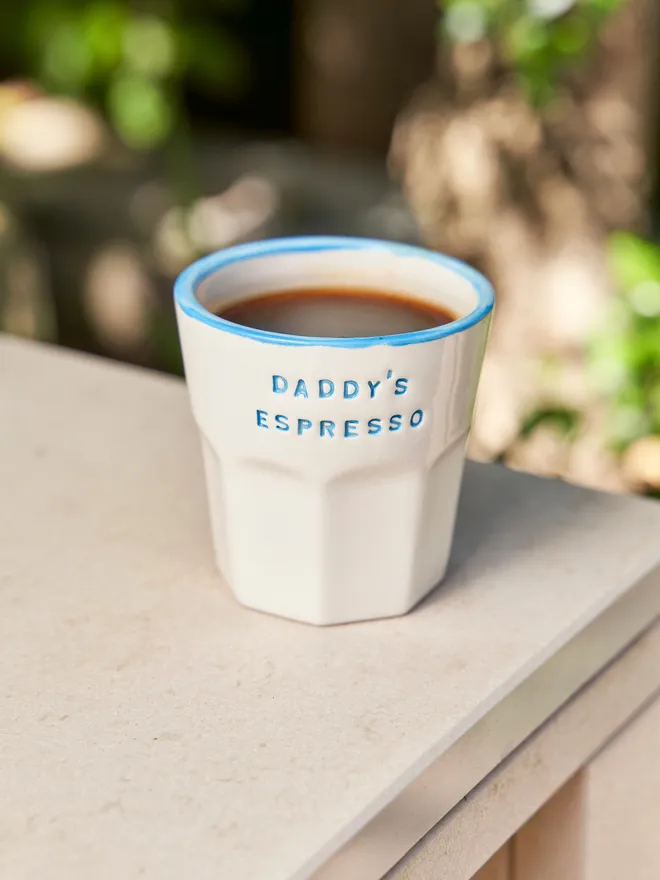 'Daddy's Espresso' in a blue font stamped on a ceramic cup with a blue rim, on a table outside