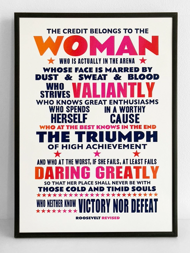 Framed print with navy blue, pink and red text - Roosevelt quote that’s been altered from the man in the arena to "The credit belongs to the woman who is actually in the arena, whose face is marred by dust and sweat and blood; who strives valiantly ...who spends herself in a worthy cause; who at the best knows in the end the triumph of high achievement, and who at the worst, if she fails, at least fails while daring greatly, so that her place shall never be with those cold and timid souls who neither know victory nor defeat." 