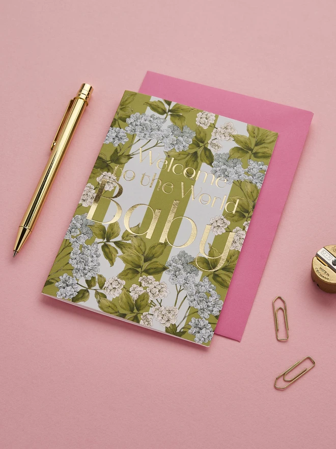 'Welcome to the World baby' card with gold foil wording and illustrated blue hydrangea flowers on a green and light blue striped base with a hot pink envelope