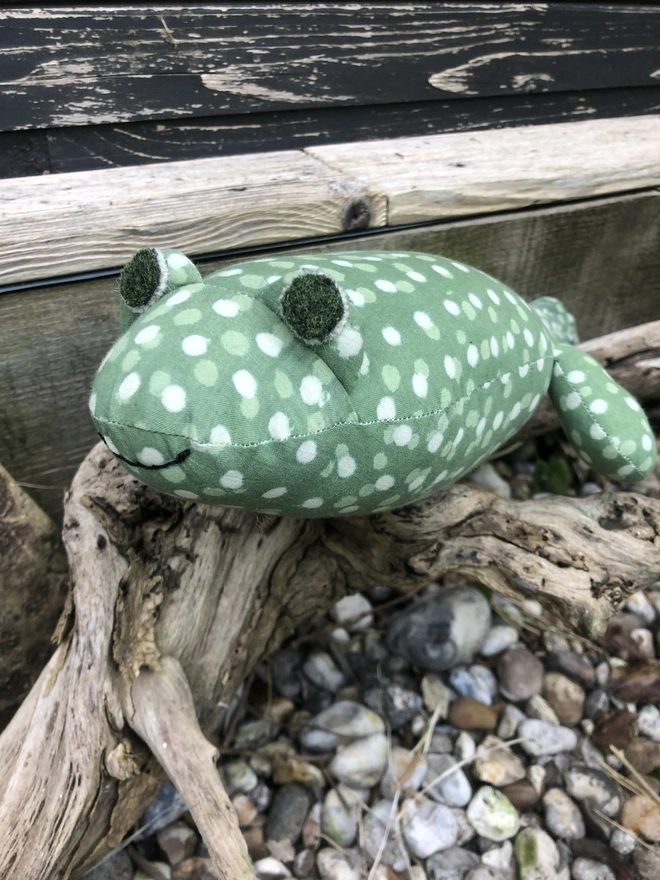 Handmade soft, fabric toy tadpole, made out of green and white spotted cotton with sewn on felt eyes, sitting on a piece of driftwood
