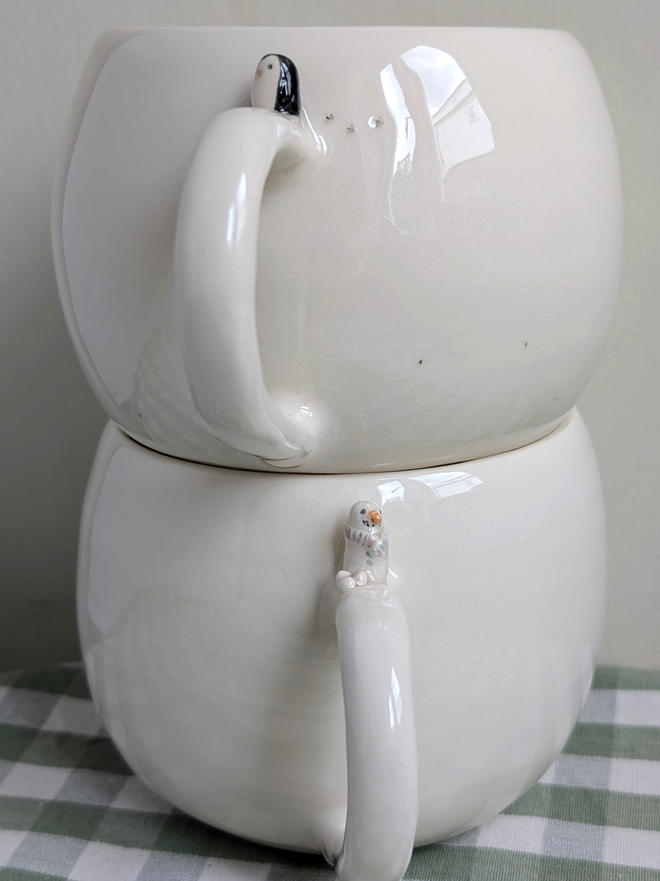 stack of 2 white cups with a tiny ceramic snowman and snowballs on the handle of one cup and a tiny penguin and bird prints on the other mug