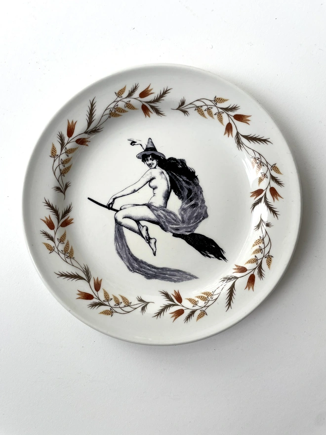 vintage plate with an ornate border, with a printed vintage illustration of a witch on a broomstick in the middle 