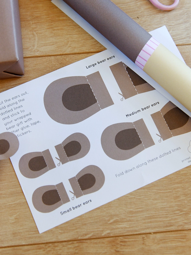 Three gifts wrapped as brown bears lay on a wooden floor beside a sheet of wrapping paper and scissors.