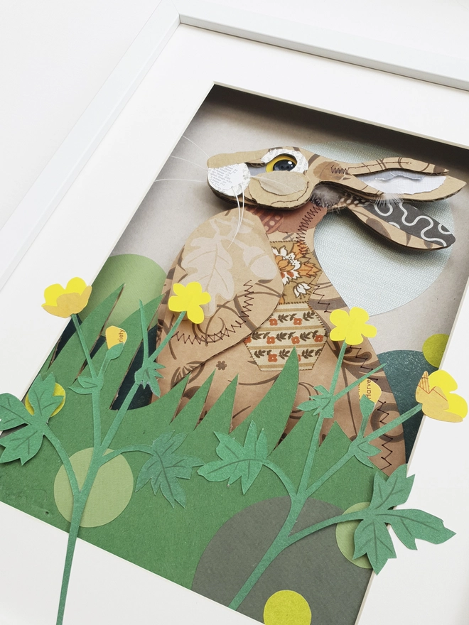 Alert upright hare nestled in long grass with buttercups in the foreground. created with layers of corrugated card to create a 3-D effect. Decorated with stitched fabric and paper.