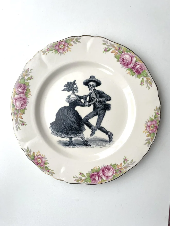 vintage plate with an ornate border, with a printed vintage illustration of two dancing day of the dead skeletons wearing a dress and a suit in the middle