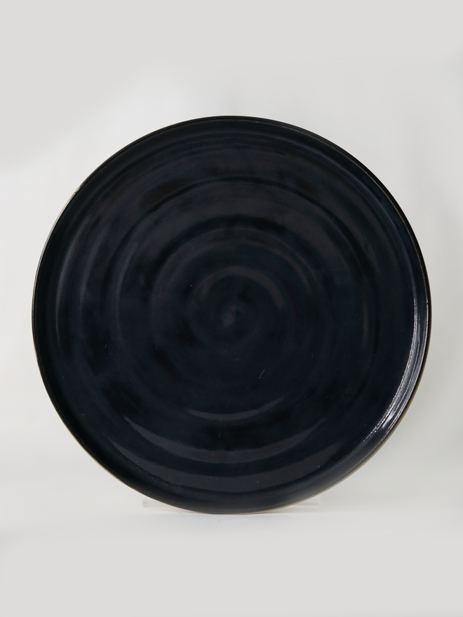 A handmade ceramic lunch plate in glossy jet black glaze, photographed from above against a plain light grey background, showing a natural concentric swirl texture from the wheel-throwing process