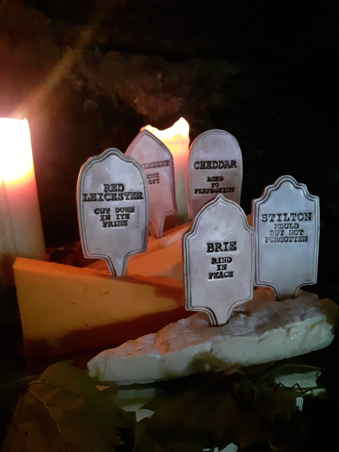Atmospheric side view of five ceramic gravestone cheese markers spread across a candlelit cheeseboard, with leafy foliage in the foreground.