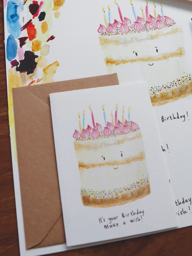 Close Up Slightly Out Of Focus Shot Of The Birthday Cake Card Sitting Along Side The Original Hand Painted Watercolour Illustration, Paintbrush and Palette