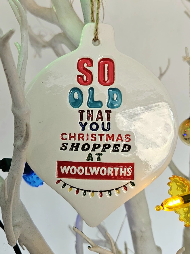 A handmade flat white bauble heart which measures 10cm by 8cm with So old that you Christmas shopped at Woolworths written on it in colourful glazes and retro fonts with a little string of colourful fairy lights stamped into the clay.