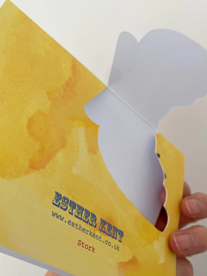 Reverse of a new baby card by Esther Kent showing a black and white stork carrying a baby in a blue checked cloth, on a yellow background