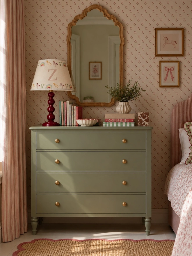 Studio Zef personalised cream unicorn lampshade featuring hand-printed unicorns, rainbows, shooting stars and the letter Z, paired with a burgundy bobbin lamp on a sage green chest of drawers in a pretty girls' bedroom with floral wallpaper and a vintage-style mirror.