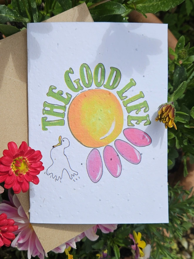 A white seed card with an illustration of a pink flower and a white bird with The Good Life written using a retro font in green. 
