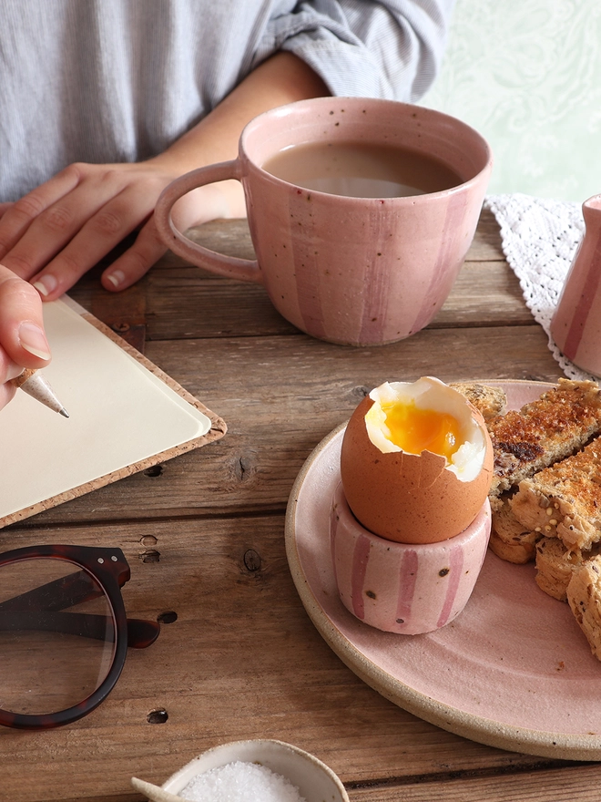 A table scene showing a Handmade Ceramic Stoneware Mug in Pink Stripe filled with coffee, a ceramic egg cup in the same style holding an egg on top of a plate of toast, a notebook and glasses, and a small ceramic dish filled with salt. The arms of a female holding a pen poised above the notebook can be seen in the background. 