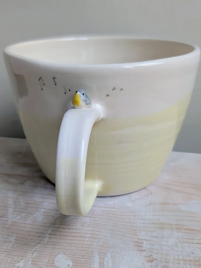 ceramic yellow bird cup with a tiny yellow and grey robin bird on the handle and musical notes and bird prints on the mug