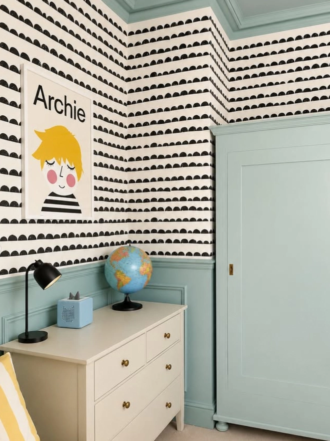 Children's room with patterned wallpaper, dresser, and globe. A personalised portrait hangs on the wall with the name Archie and an image of an illustrated boy with yellow hair