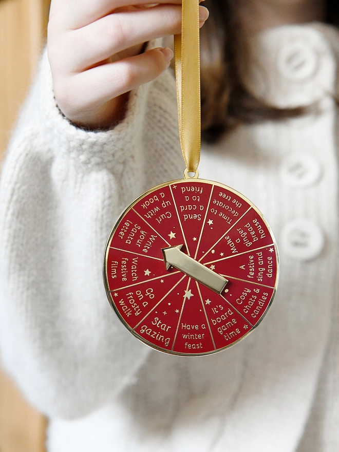 A deep red and gold Christmas decoration is being held by a gold ribbon. It has 12 segments, each one with a different Christmas activity idea, and a golden arrow in the centre.